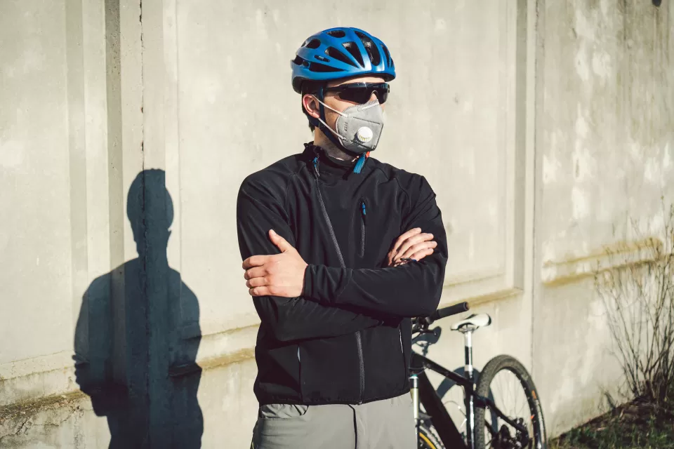 Male cyclists wearing helmet, sunglasses, and N95 mask stands with arms crossed in front of his bike